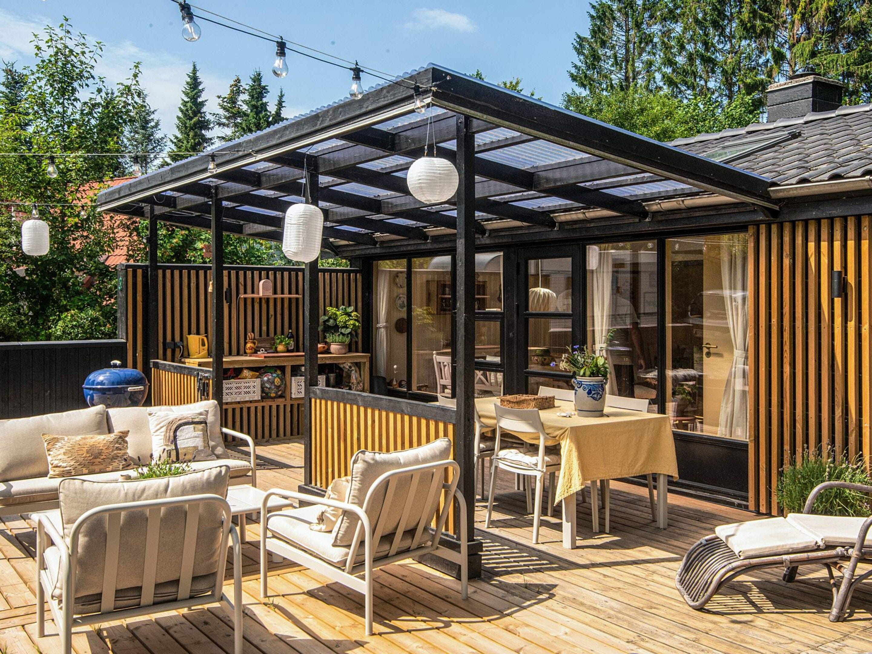 A covered outdoor patio with seating and a table, surrounded by wooden fences and plants.