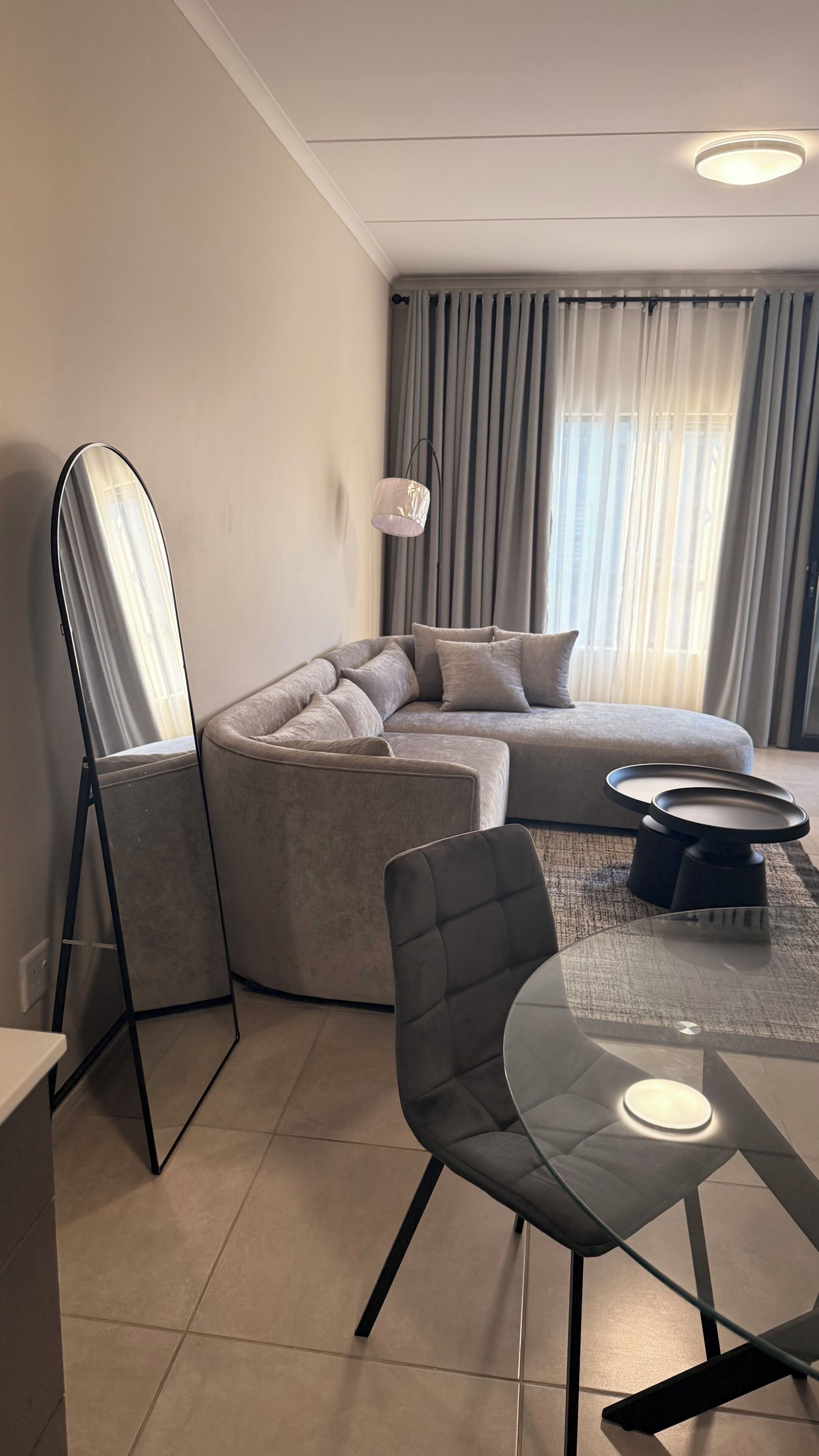 A modern living room with a sectional sofa, a glass coffee table, a chair, a floor lamp, and a large mirror.