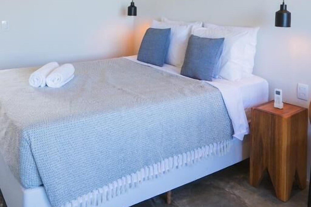 A bedroom with a bed, bedside tables, a wall-mounted air conditioner, and framed artwork.
