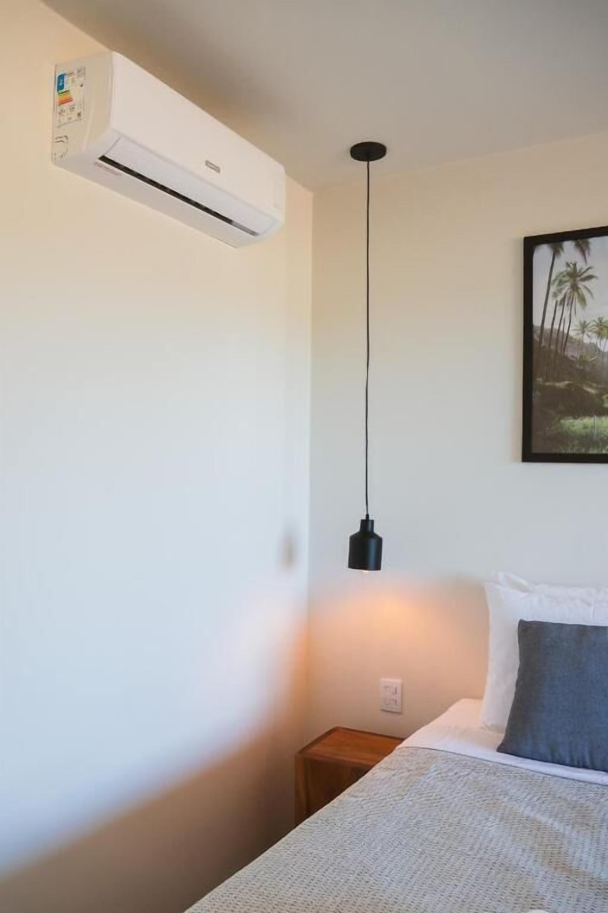 A bedroom with a bed, a nightstand, an air conditioning unit, a hanging light fixture, and a framed picture on the wall.