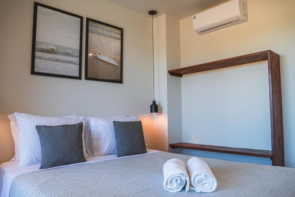 A hotel room with a bed, two pillows, a rolled towel, an air conditioner, and two framed pictures on the wall.