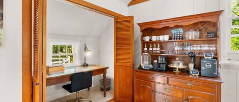 A cozy room with a wooden desk, a coffee station, and a view of greenery outside.