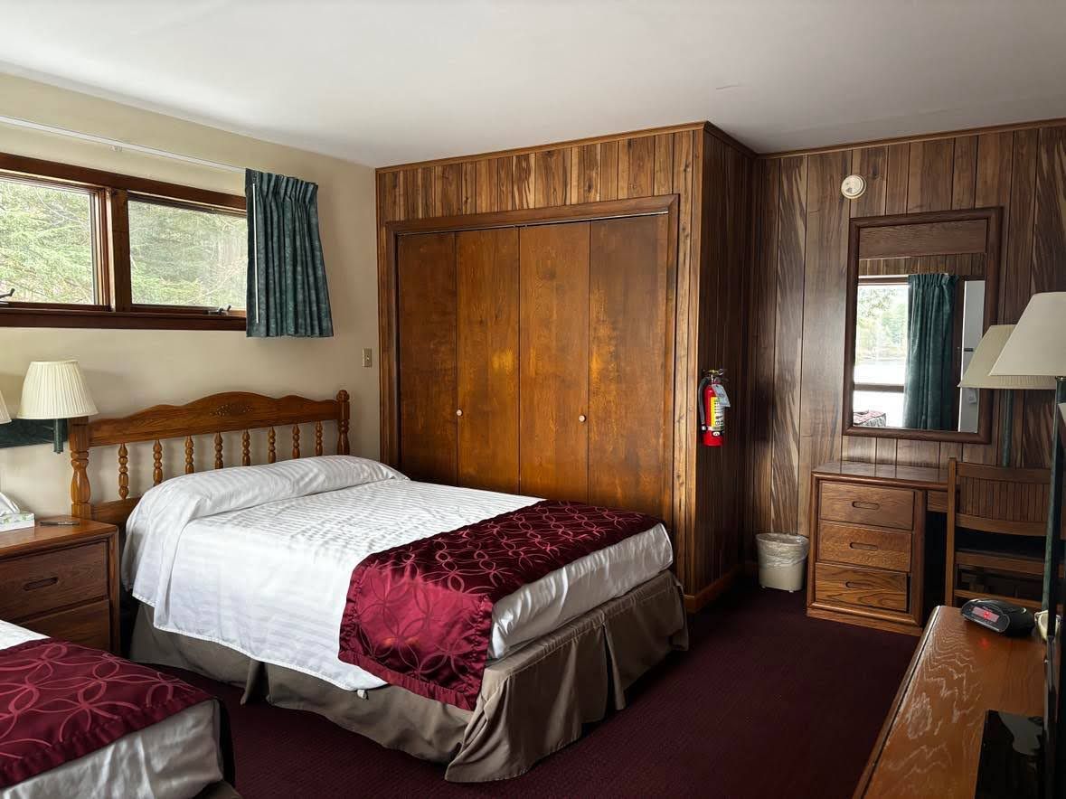 Un dormitorio con un armario de madera, una cama con cubrecama rojo y blanco, una mesita de noche con lámpara y una ventana con cortinas.