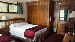 Een slaapkamer met een houten garderobe, een bed met een rood-witte spreidek, een nachtkastje met een lamp en een raam met gordijnen.