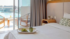 Ein Hotelzimmer mit Bett, Esstisch mit Obstkorb und Tasse, Stuhl, Blick auf ein Gewässer und Balkon mit Sitzgelegenheit.