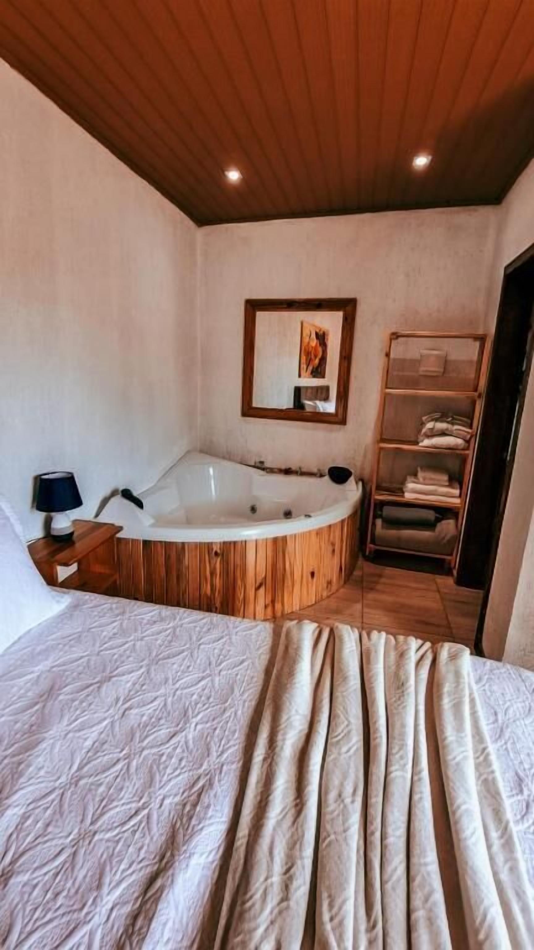 A room with a large bed, a wooden bathtub, a wooden ceiling, a shelf with towels, and a framed picture on the wall.
