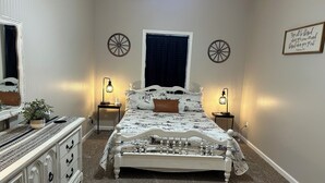 A bedroom with a bed, two bedside tables, a dresser, a mirror, a window with curtains, and wall decorations.