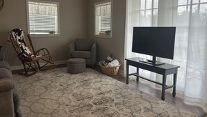 A living room with a TV, two windows with blinds, a rocking chair, a grey armchair, and a patterned rug.