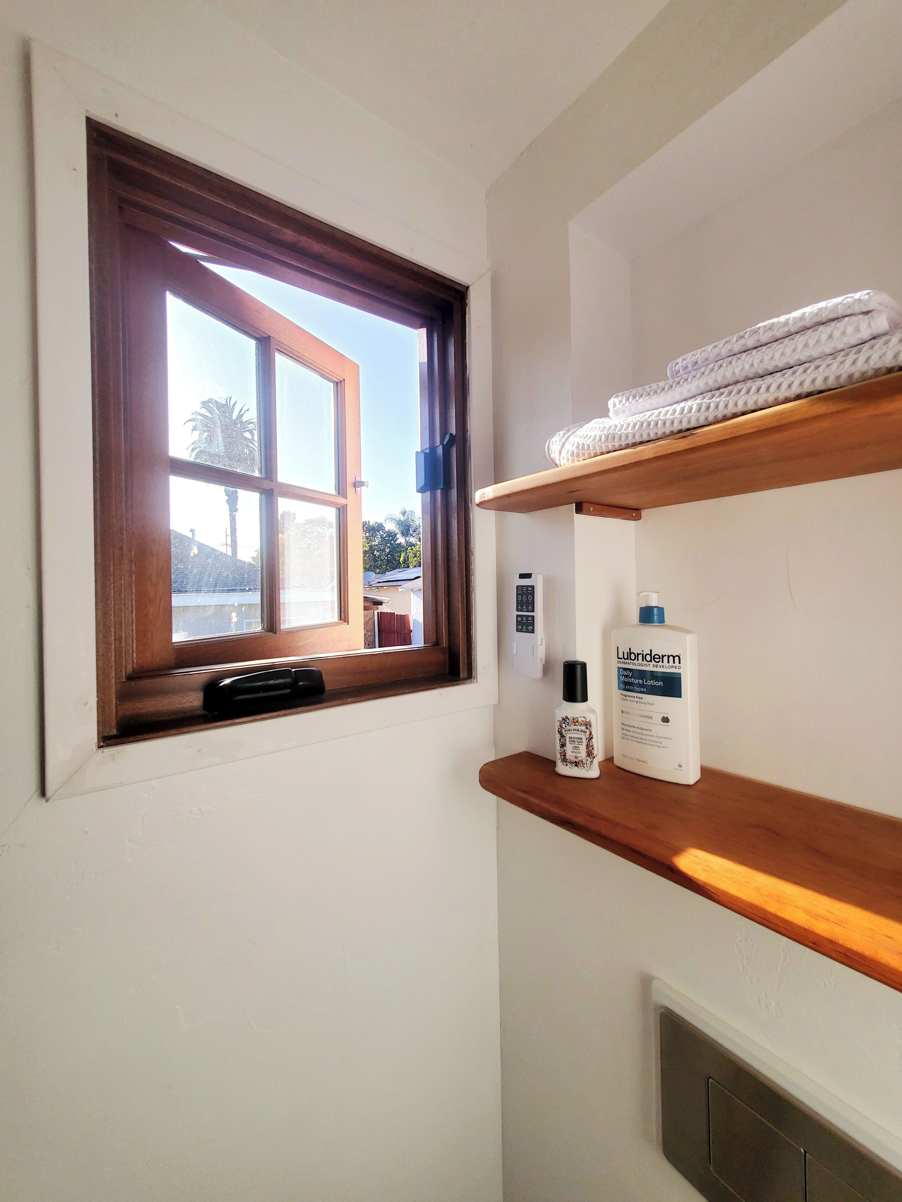 A bathroom with a window, a thermostat, a shelf with towels, and a bottle of Lubriderm lotion.