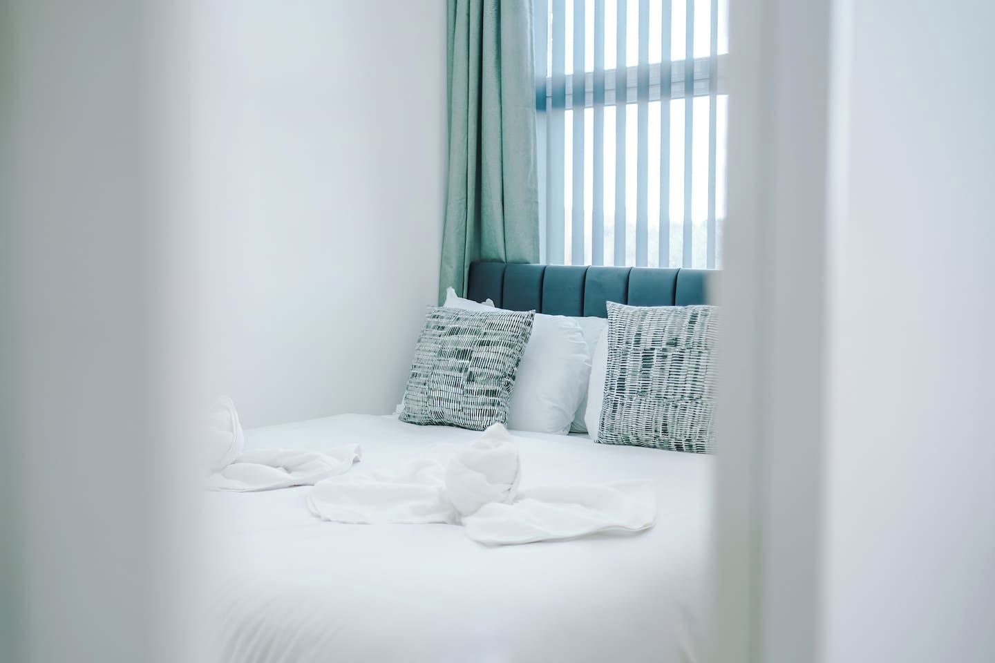 Ein ordentlich bezogenes Bett mit weißen Leinen und zwei gemusterten Kissen, ein Fenster mit vertikalen Jalousien und ein türkisblaues Kopfteil.