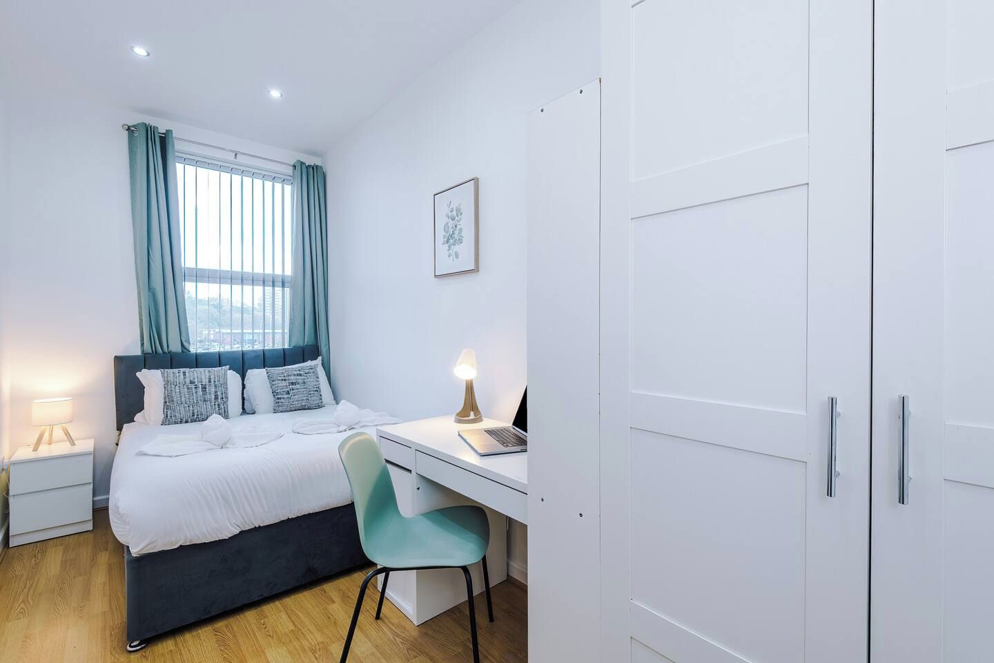 Ein Schlafzimmer mit Bett, Nachttisch, Lampe, Schreibtisch mit Laptop, Stuhl und Kleiderschrank.