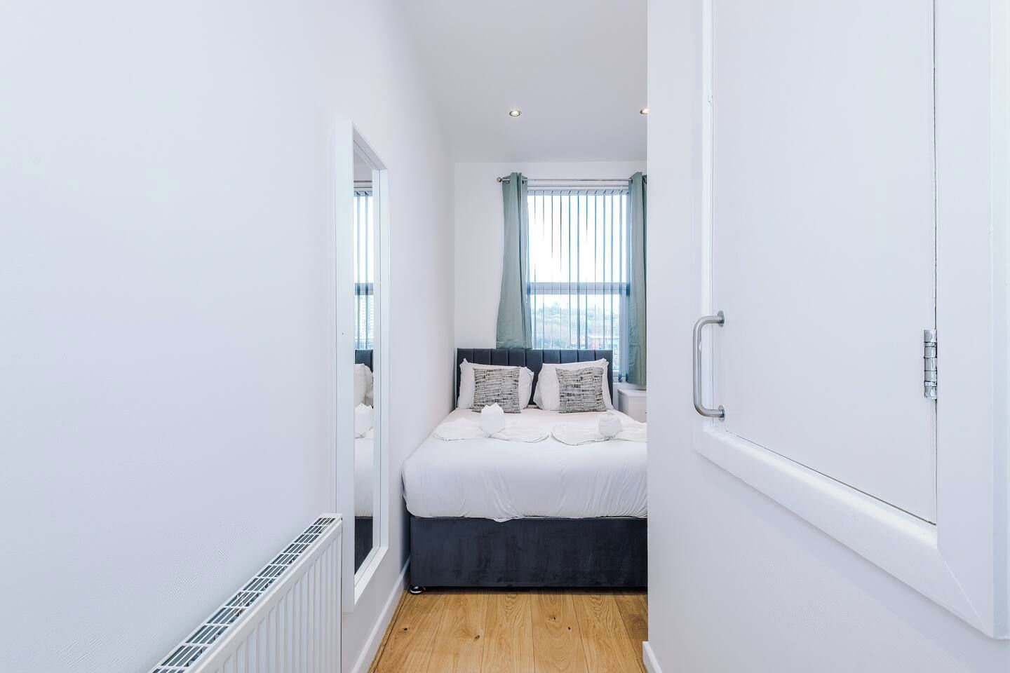 Ein schmaler Flur führt zu einem Schlafzimmer mit einem Bett, einem Fenster mit Vorhängen und einem Heizkörper.