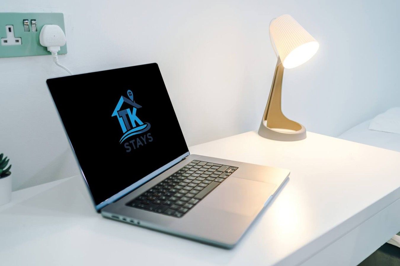 Ein Laptop auf einem weißen Schreibtisch, mit einem Logo auf dem Bildschirm, einer Lampe und einer Steckdose.