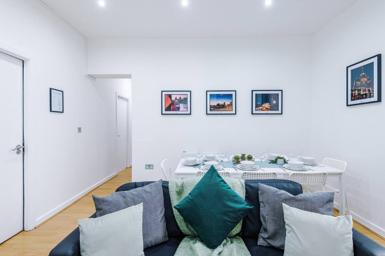 Ein moderner Essbereich mit einem weißen Tisch für vier Personen, Stadtbilder an der Wand und eine Sitzecke mit vielen Kissen im Vordergrund.