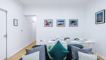 Ein moderner Essbereich mit einem weißen Tisch für vier Personen, Stadtbilder an der Wand und eine Sitzecke mit vielen Kissen im Vordergrund.