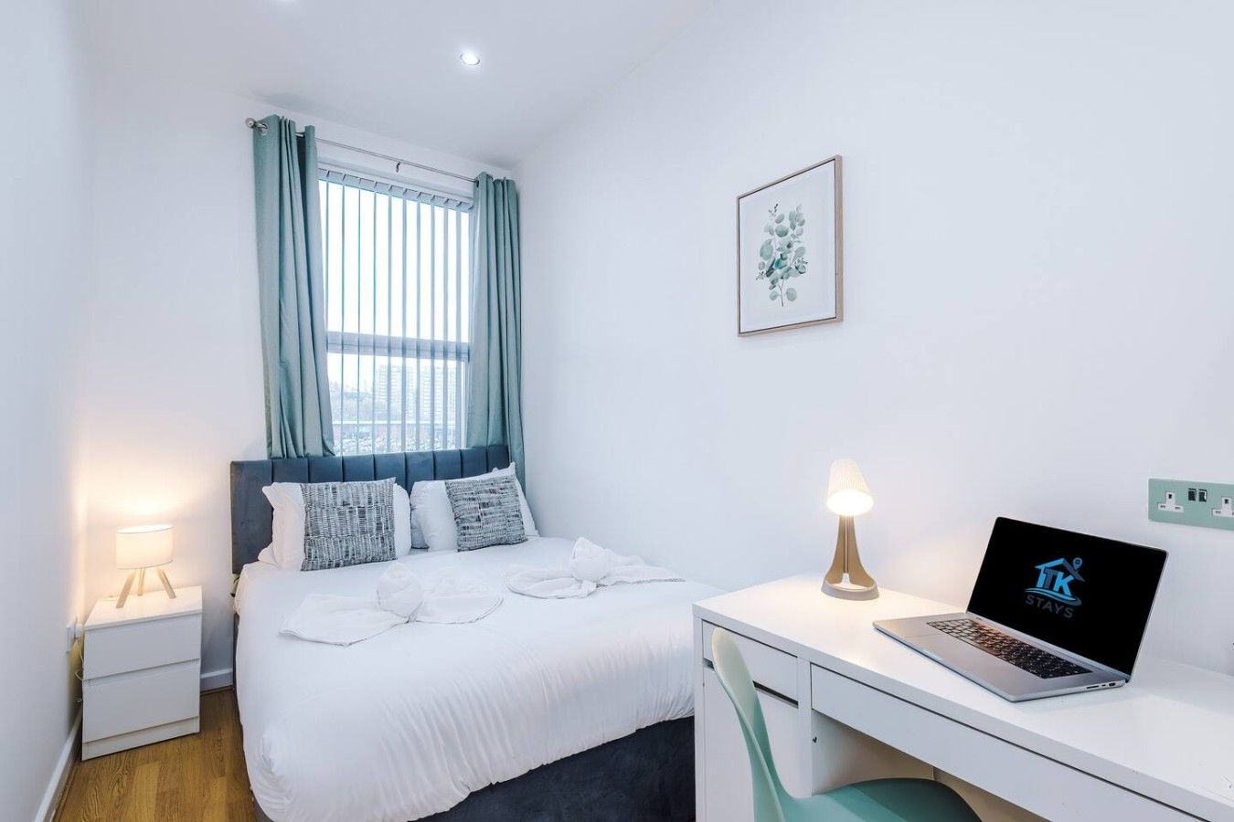 Ein Schlafzimmer mit einem Bett, einem Schreibtisch mit Laptop, einer Nachttischlampe und einem Fenster mit Vorhängen.