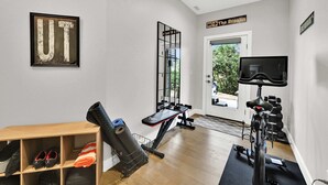 Ein Fitnessraum mit Trainingsgeräten, einer Bank, einem an der Wand montierten Fernseher und einer zur Außenseite führenden Tür.