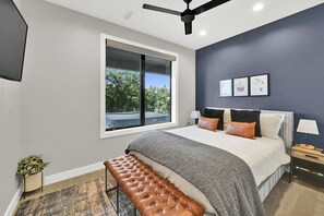 A modern bedroom with a large bed, a bench, a ceiling fan, and a window with a view of trees.
