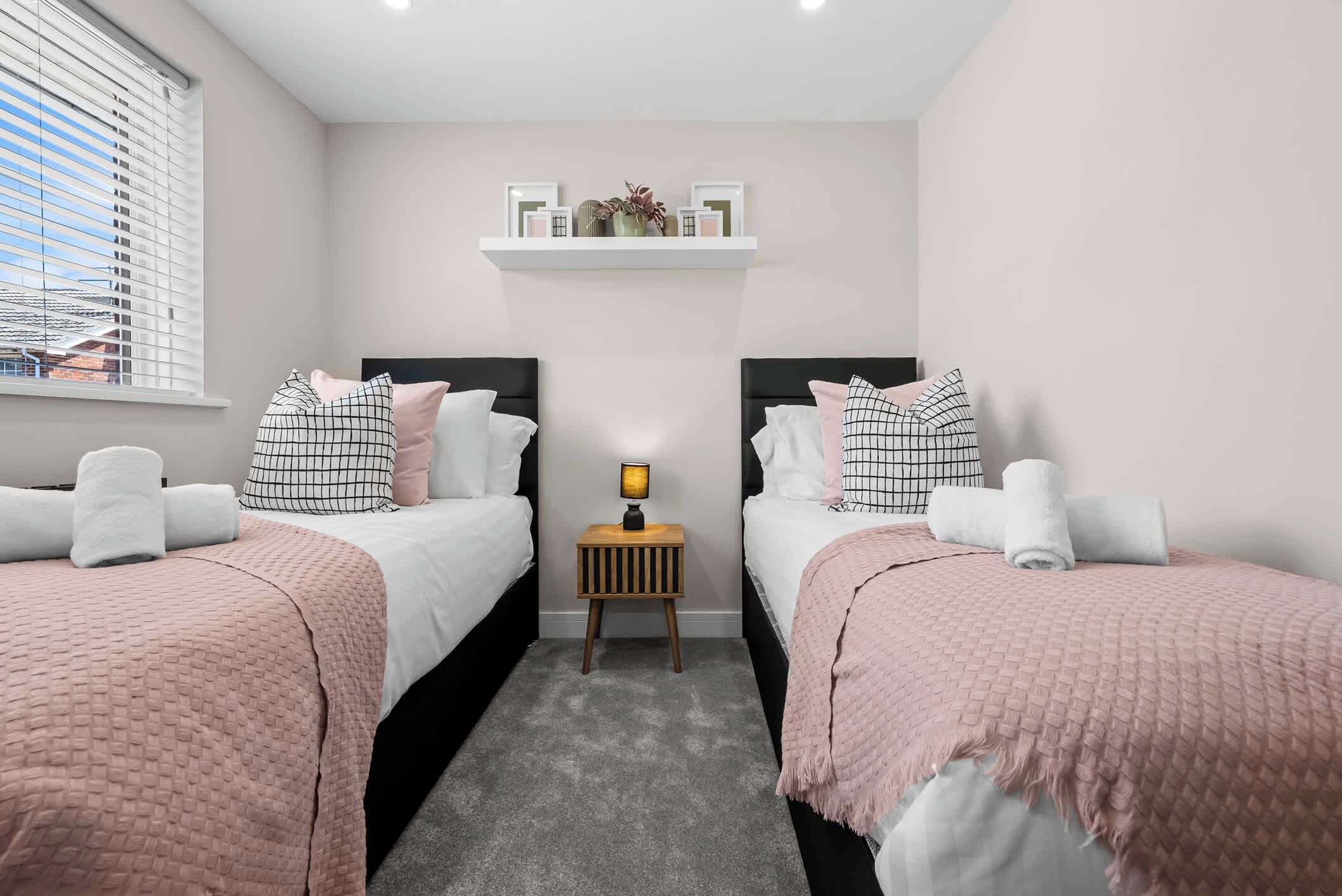 A bedroom with two beds, a nightstand with a lamp, a shelf above the beds, and a window with blinds.