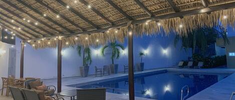 An indoor pool area with a thatched roof, lounge chairs, and potted plants.