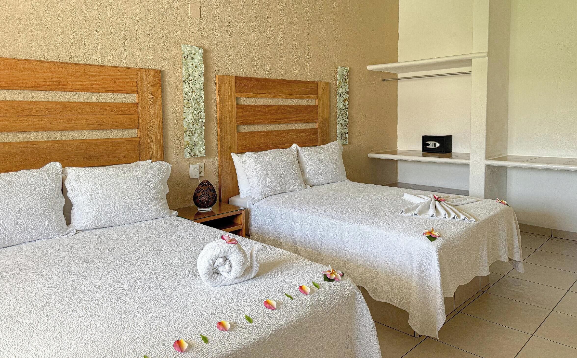 A hotel room with two beds, each with white linens and decorative pillows. There is a wooden headboard, a nightstand with a lamp, and a shelf with a small item on it.