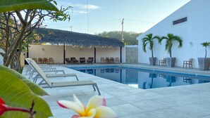A swimming pool with lounge chairs and a thatched-roof structure, surrounded by palm trees and a white building. - Hotel y Condominios Jaroje (Crucecita)