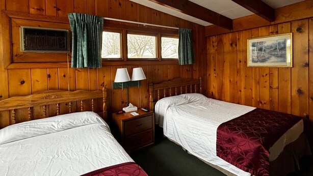 Una stanza con rivestimento in legno, due letti, un comodino con lampade, una finestra con tende e un quadro appeso al muro.