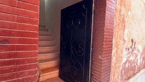 Ein Eingang aus roten Ziegeln mit einer schwarzen, verzierten Tür, einem Korbuschel darüber und einer Treppe, die hineinführt.