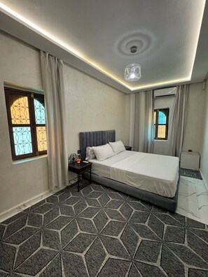 A hotel room with a bed, a nightstand, a lamp, a window with curtains, and a geometric-patterned carpet. - Stylish 2-Bedroom Apartment in Marrakech Near Jemaa el-Fnaa (Marrakesh)