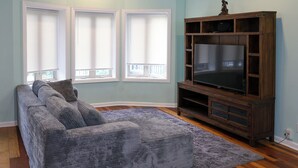 A living room with a grey sectional sofa, a flat-screen TV on a wooden stand, and a window with blinds. - Spacious 5-bedroom house in Chicago with AC Near Obama Center & UChicago (Chicago)