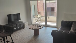 A living room with a sofa, a TV, a table, and chairs. There is a balcony with a view of a building. - Premium Luxury Apartment (Dunkerque)