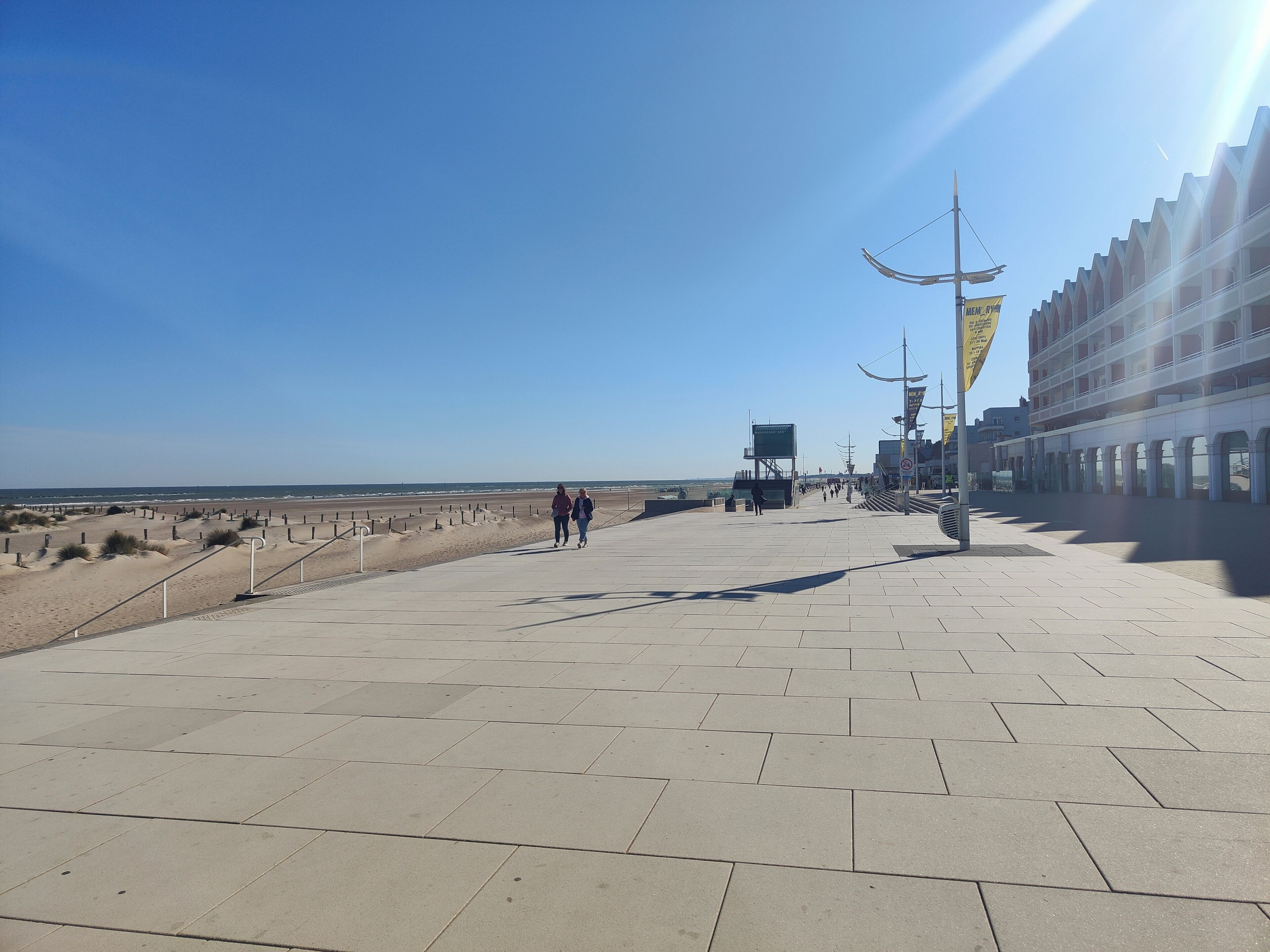 Un paseo marítimo pavimentado con una playa al fondo, un edificio con ventanas arqueadas y un cielo despejado.