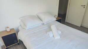 A neatly made bed with white linens, a rolled towel, and a bedside table with a lamp and a power outlet. - Premium Luxury Apartment (Dunkerque)