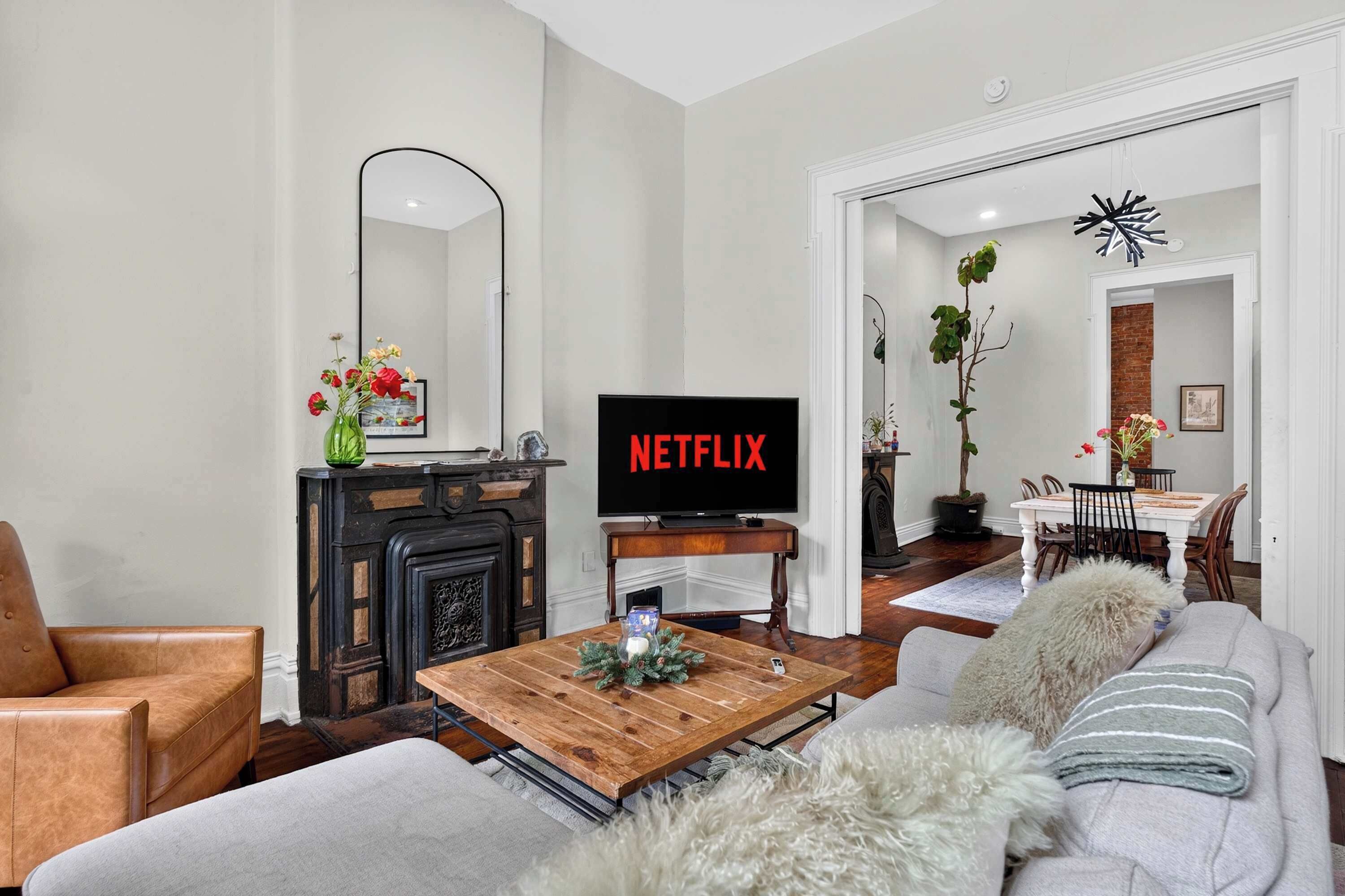 A living room with a fireplace, a television displaying 'NETFLIX', and a dining area visible through an open doorway.