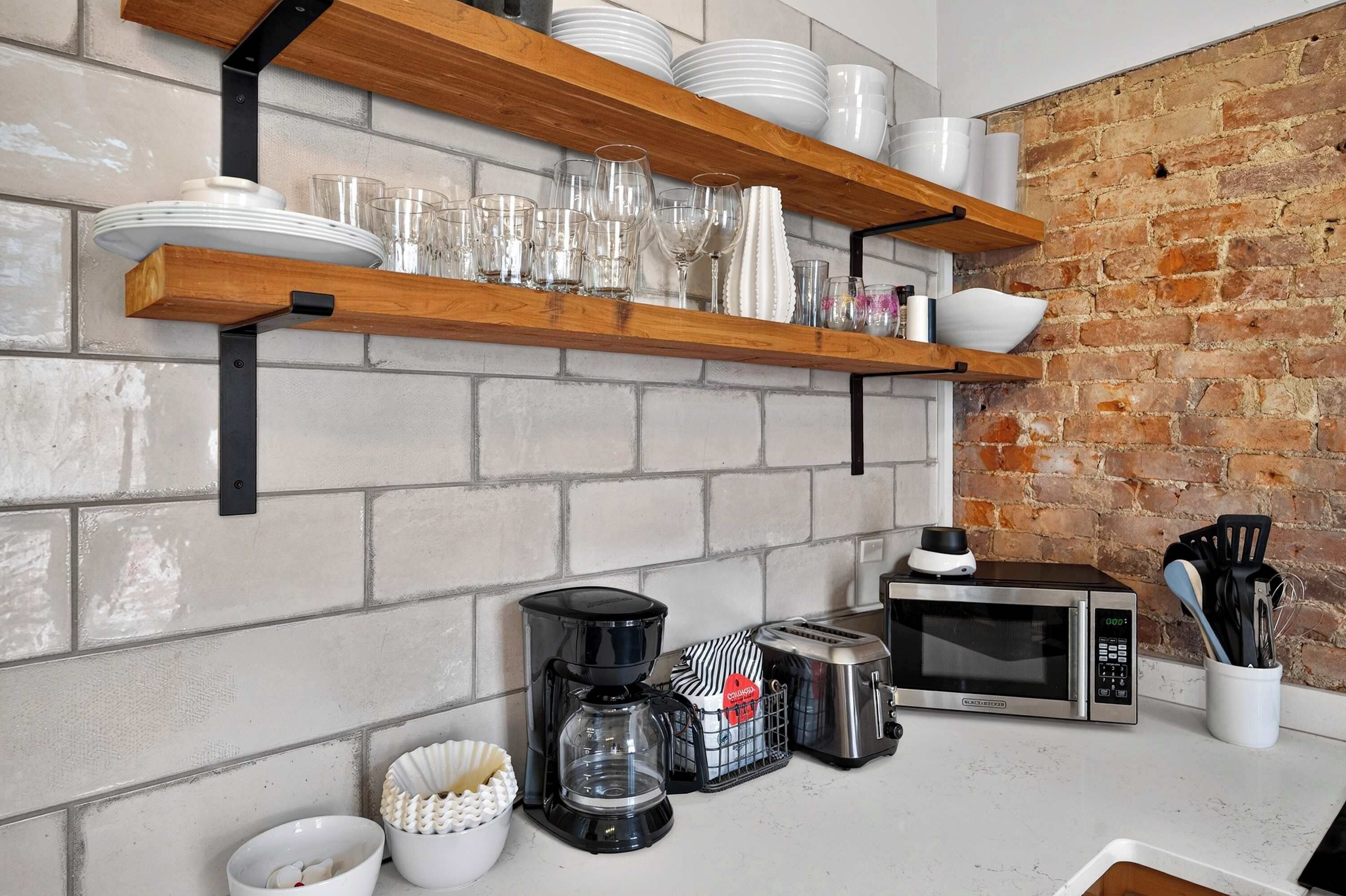 Una cocina con paredes de ladrillo blanco, estantes de madera con platos y vasos, una cafetera, un tostador y un microondas.