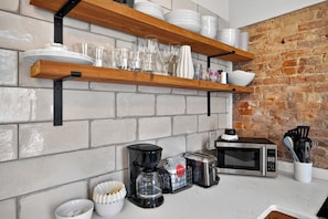 A kitchen with white brick walls, wooden shelves holding plates and glasses, a coffee maker, a toaster, and a microwave. - Relaxing home w/ poker lounge & spacious backyard! (Louisville)