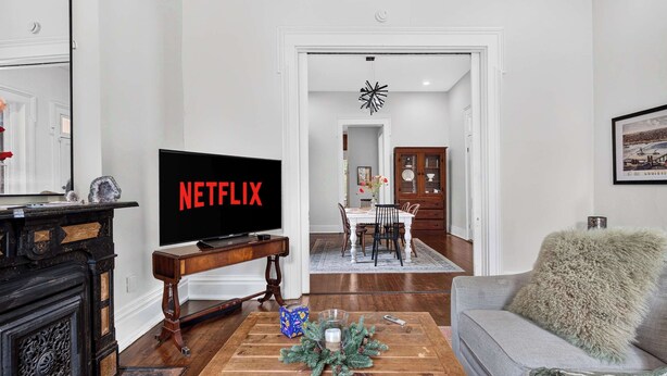 Ein Wohnzimmer mit einem Fernseher, der 'NETFLIX' zeigt, einem Holztisch mit einer Dekoration, einem grauen Sofa und einem Flur, der in einen Essbereich führt.