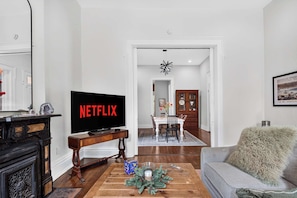 A living room with a television displaying 'NETFLIX,' a wooden coffee table with a decorative item, a grey sofa, and a hallway leading to a dining area. - Relaxing home w/ poker lounge & spacious backyard! (Louisville)