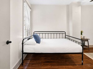 A single bed with a metal frame, white bedding, and a blue pillow in a room with wooden flooring and a window with blinds. - Relaxing home w/ poker lounge & spacious backyard! (Louisville)