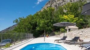 En swimmingpool med solstolar och ett parasoll, omgiven av en stenmur och grönska.