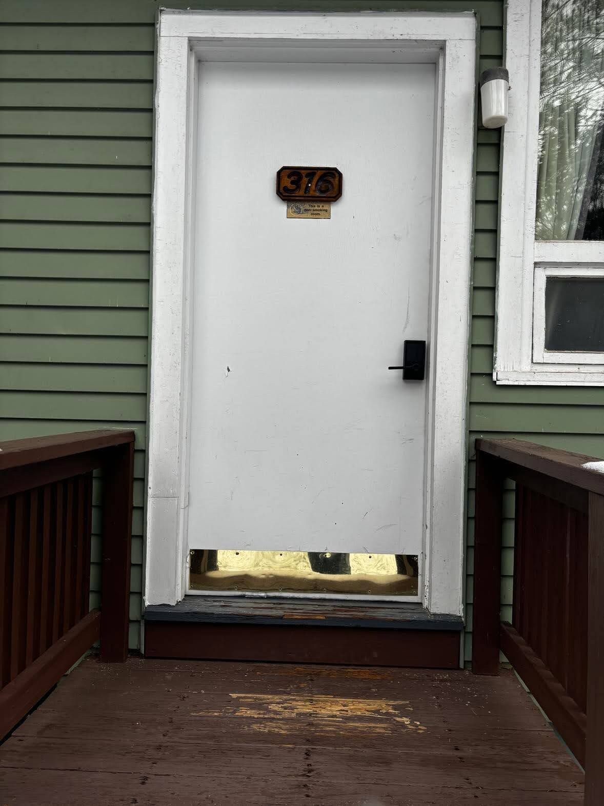 A white door with a number plate, a small window to the right, and a wooden porch with a metal step leading up to it.