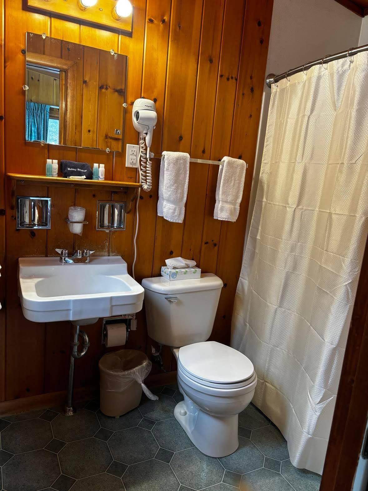 Un baño con pared de madera, un lavabo blanco, un inodoro, una cortina de ducha y un secador de pelo.