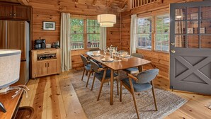 Interno di una baita in legno con tavolo da pranzo apparecchiato per quattro persone, angolo cottura e una grande finestra con tende.