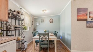 Comedor con mesa de madera, sillas y zona de cocina con lavavajillas y estanterías.