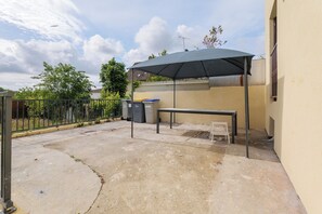 An outdoor patio with a table and bench, a covered area, and trash bins. - Villa LAYRA | A Luxury Haven of Peace (Goussainville)