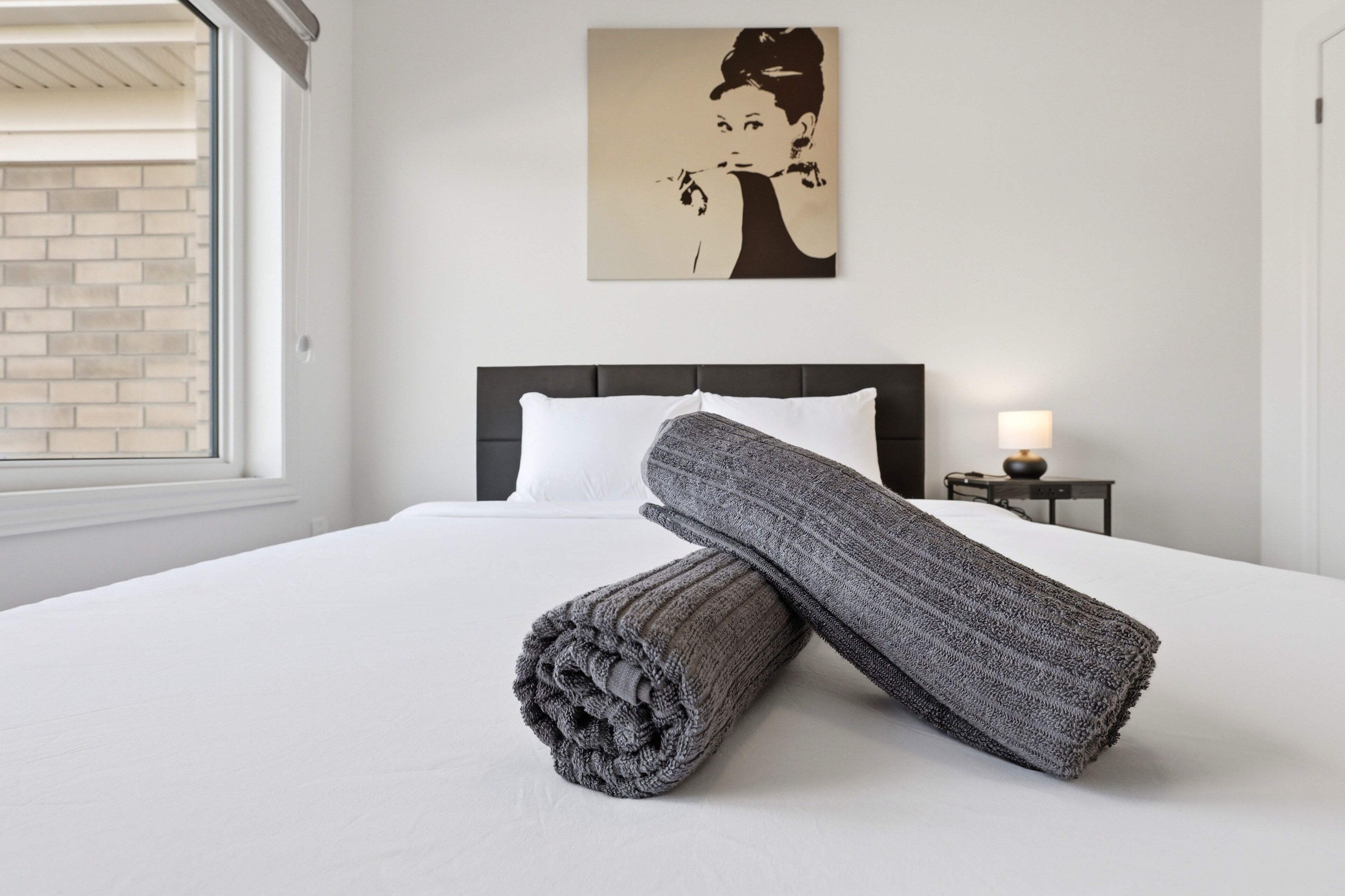 A neatly made bed with rolled towels, a black headboard, a wall-mounted picture, and a small bedside table with a lamp.