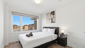 A bedroom with a large window, a bed, a bedside table with a lamp, and a wall-mounted artwork.