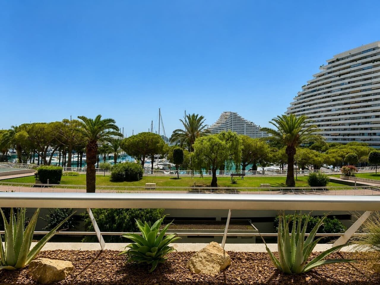 A modern urban park with palm trees, a water feature, and a large building in the background.