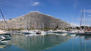 Marina con varios veleros amarrados frente a un amplio hotel moderno de diseño escalonado.