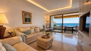 Sala de estar con sofá, reposapiés y televisor. Se ve el océano a través de amplios ventanales.